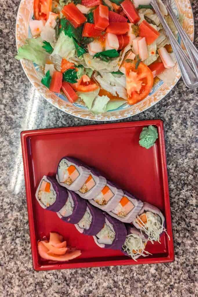 Overhead shot of two dishes. Topmost is a salad of iceberg lettuce, tomato, and vegan salmon. Bottom is sushi wrapped in mashed taro and purple sweet potato instead of rice