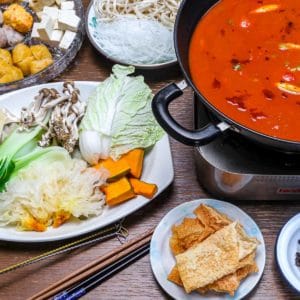 Table spread with various hot pot ingredients (tofu, vegan shrimpballs, vegetables, fried yuba, noodles), and tomato hotpot base