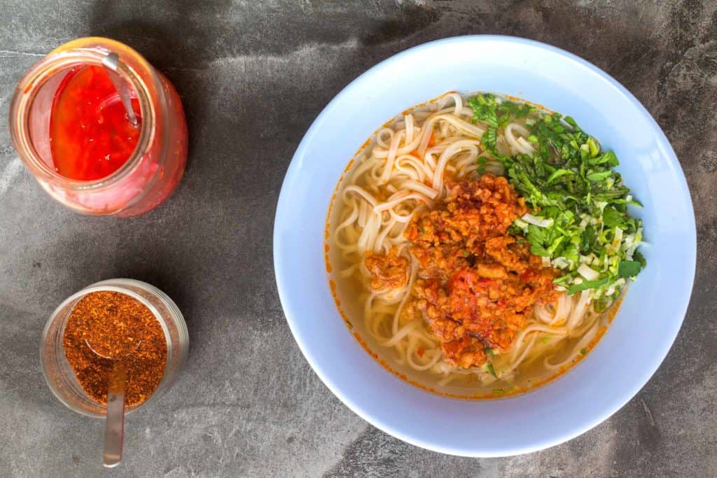 bowl of noodles with chilli and tomato ragu on top, plus coriander and spring onion. To the left of the bowl are jars of toasted chilli powder and chilli vinegar.