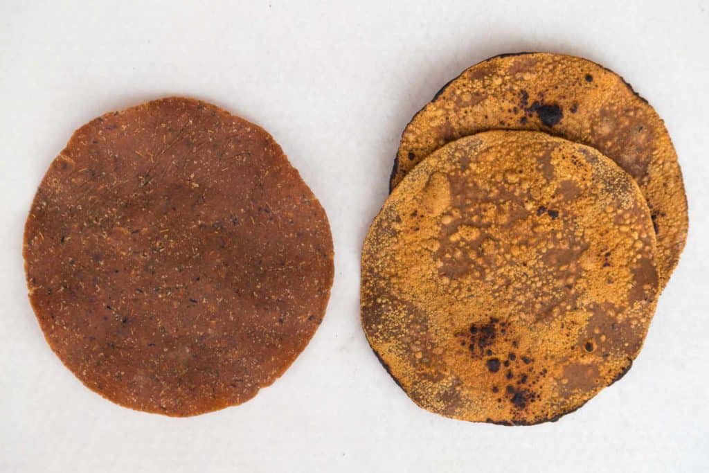 Round, brown discs of a dried fermented soybean product called tua nao. The left piece is uncooked and the right piece is partially grilled.