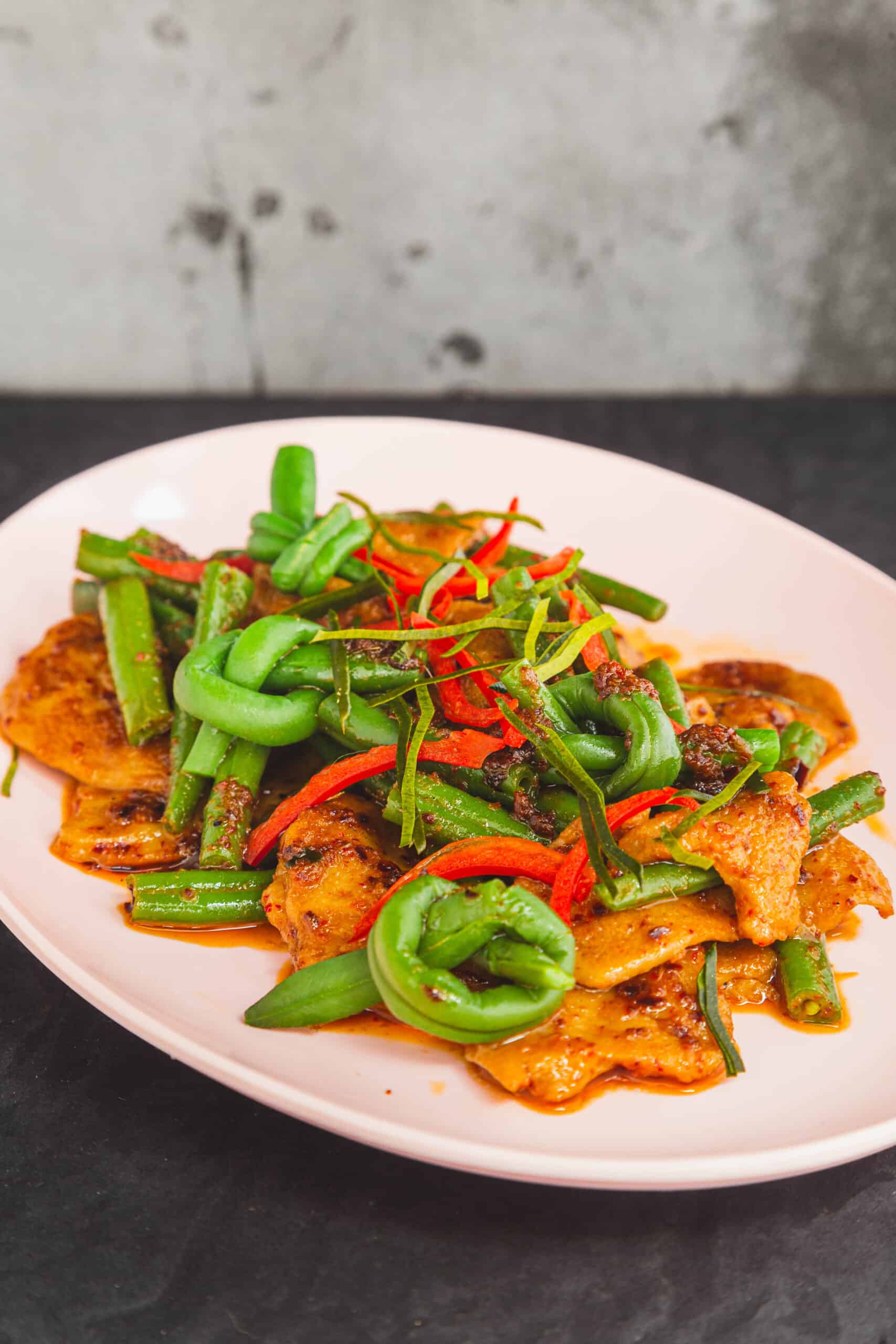 light pink oval melamine dish on a dark background. Plate contains stir fried seitan with green beans in red curry paste (vegan pad prik king)