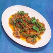 photo of a light pink oval melamine dish on a blue background (painted wood). The dish sets at a 45 degree angle. The plate contains stir fried seitan with red curry paste and green beans. Some of the green beans are tied into knots.