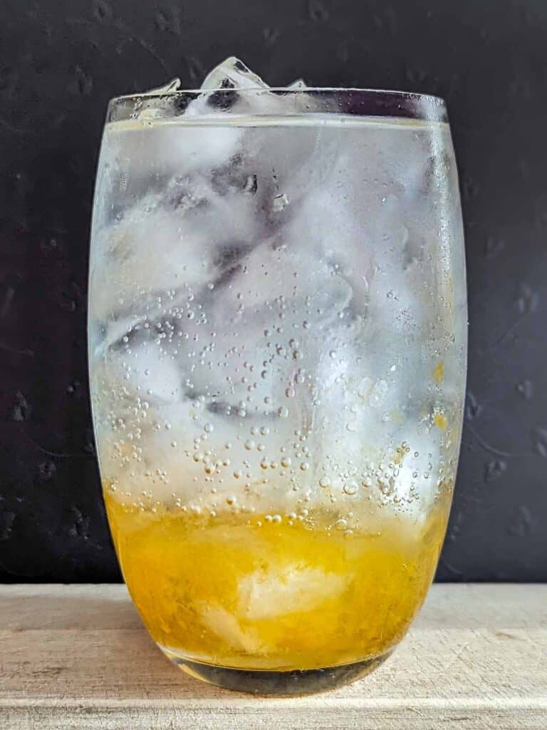 starting with the background: top ⅘ is black. bottom ⅕ is light wood, on which a glass of liquid sits. The glass is full to the brim with ice. The bottom ⅓ of the glass is amber coloured (salted plum), while the remaining space in the glass is clear soda water. Some bubbles are formed on the inside of the glass and drops of moisture on the outside.