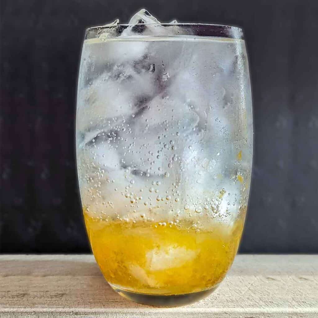 starting with the background: top 4/5 is black. bottom 1/5 is light wood, on which a glass of liquid sits. The glass is full to the brim with ice. The bottom 1/3 of the glass is amber coloured (salted plum), while the remaining space in the glass is clear soda water. Some bubbles are formed on the inside of the glass and drops of moisture on the outside.