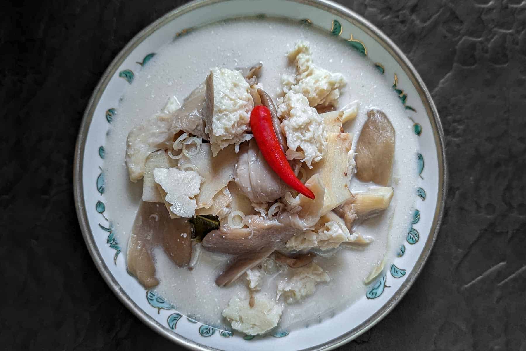 Black background with a bowl of tom kha soup on top. The bowl is shallow and features a silver rim and some blue-green pattern deeper in the bowl. The bowl contains white coconut based soup, with vegan chicken and oyster mushrooms piled up in the middle. There is a single small red chilli on top.
