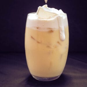 dark background with slightly curved glass filled 5/6 of the way with beige-yellow liquid (corn milk), topped with a creamy cheese foam (white). The foam is messily dripping down the side of the glass.