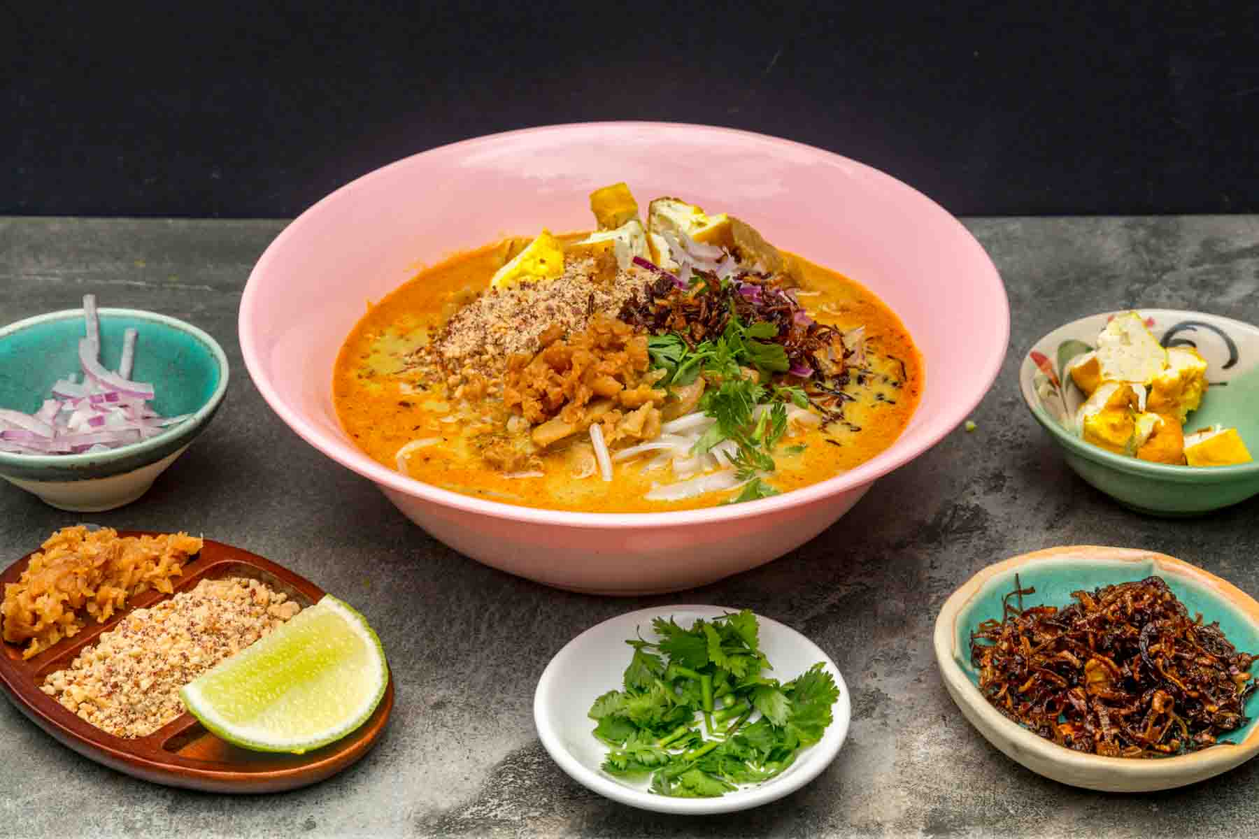 pink melamine bowl filled with orange coloured curry. Some noodles are visible underneath the heaped toppings comprised of cubes of fried tofu, ground peanut, preserved radish, fried shallots, coriander, and sliced shallot.