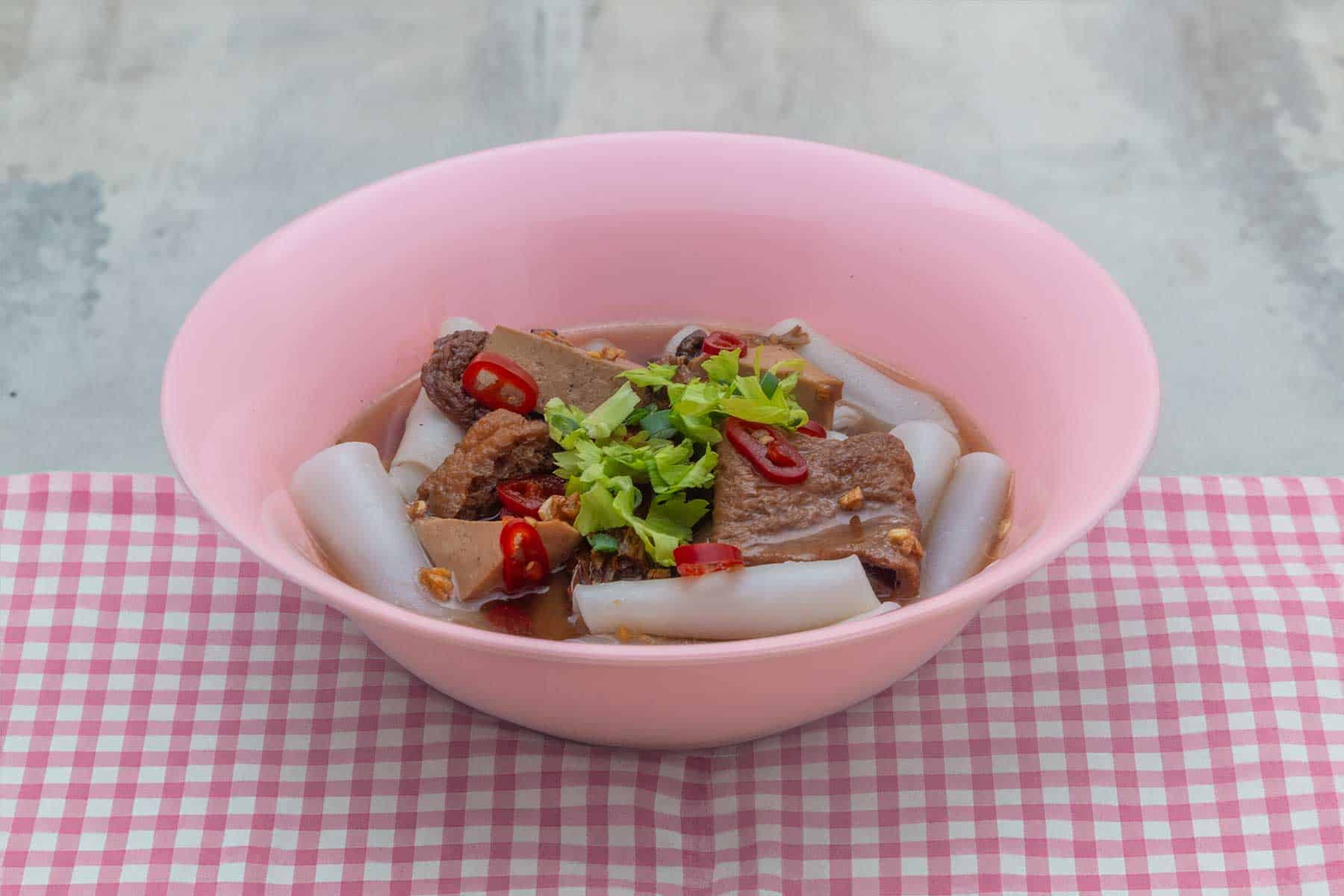 Vegan kway jap nam kon - rolled rice noodles in cloudy broth with mock meats, in a pink bowl, sitting on a red checkered tablecloth