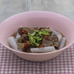 Rolled rice noodles in cloudy broth with mock meats, in a pink bowl, sitting on a red checkered tablecloth