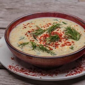 cream coloured soup in a brown bowl that is sitting on a white dish on a wooden background. The soup is garnished with sprigs of green dill and bright red aleppo pepper.