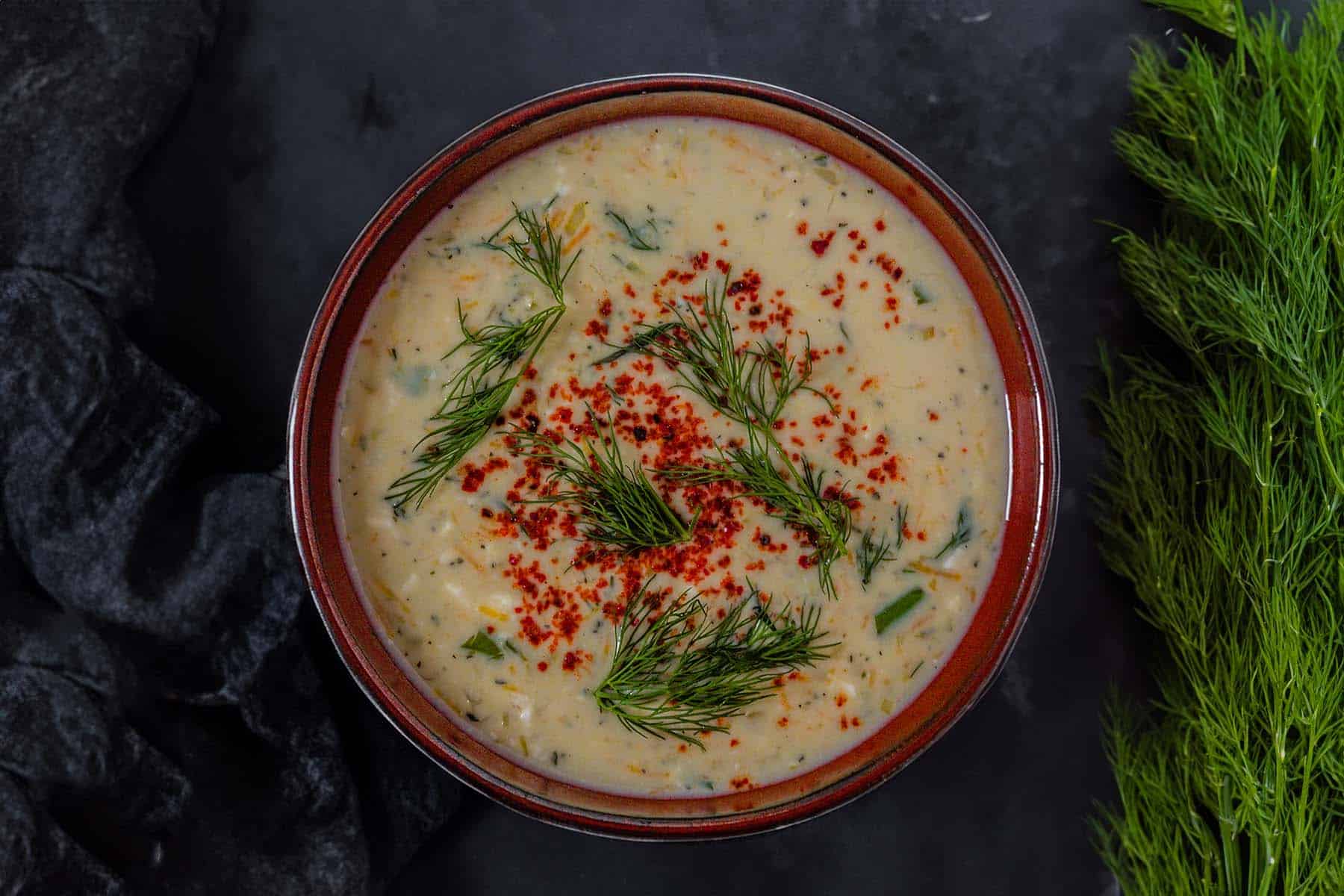 cream coloured vegan yoghurt soup in a brown bowl on a black background. The soup is garnished with sprigs of green dill and bright red aleppo pepper. A large bundle of dill sits on the side of the bowl.