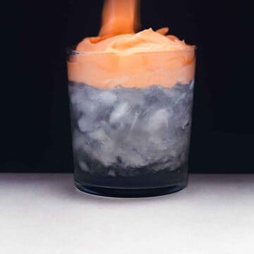 Short ice-filled glass on a white surface against a black background. The glass contains 75% clear liquid (coconut water) and is topped with bright orange whipped Thai tea.